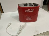 Red Coca-Cola hotdog and bun maker with two slots and a power cord on a white surface.