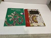 Two adult colouring books placed side by side on a white surface. Left book: green cover titled 'Nirvana Colouring for Everyone' with Om symbol artwork. Right book: 'The Art of Indian Mandalas' with intricate mandala cover and red spine.