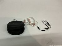 Photo showing Bluetooth earphones with white cords and rose gold earbuds, a small black zippered carrying case, and a short black dual audio cord with 3.5mm connectors on a white surface.