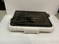 Electric griddle with black non-stick cooking surface, white base, temperature control dial, oil tray attached to side, and power cord with two-prong plug coiled on top.