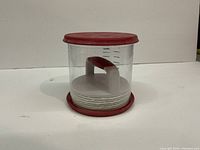 Two plastic hamburger presses stacked, with red lids and bases, clear sides with volume markings, and white handles for pressing patties.