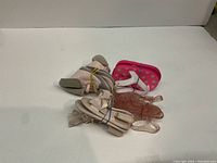 Four infant footwear items on white background. Three are baby sandals in pink, beige, and clear pink, and one is a pair of soft gray infant shoes with white/beige stripes.