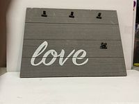 Front view of the wooden picture frame painted gray with the word 'love' in white cursive. Shows four black metal clips attached for holding photos.