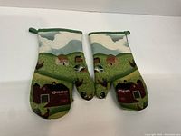 Front side of both oven mitts showing landscape farmhouse pattern with barns and animals