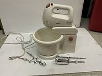 Philips 5 speed hand mixer sitting on matching stand with mixing bowl. Whisk and dough hook attachments spread around base.
