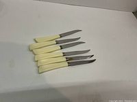 Six serrated steak knives with cream-colored handles laid side by side on a white background.