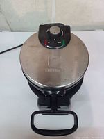Top view of closed Chefman waffle maker showing stainless steel surface, control knob with red and green indicator lights, and handle.