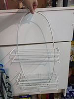 White wire shower caddy hanging against a white cabinet door, showing two shelves and a hook for hanging.