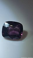 Cushion cut gemstone with deep purple to pink coloration on light background, showing facets with some internal reflection and good transparency.