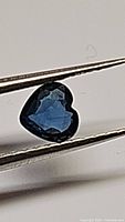 Close-up image of a heart-shaped blue sapphire held by tweezers, showing strong blue color and faceting.