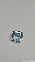 Close-up image of a square emerald cut light lagoon blue aquamarine gemstone showing clarity and cut details on a gray background.