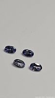 Four oval faceted blue-purple tanzanite gemstones displayed on a neutral background, showing the color and cut of each stone.