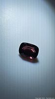 Close-up photo of the 1.07ct deep purple-red spinel gemstone on a white background, showing its cushion cut and color under natural light.