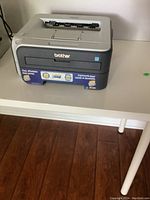 Front left perspective of the gray Brother HL-2140 laser printer showing the paper output area on top and branding on front.