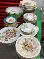 Wide view of assorted bone china plates and bowls arranged on a green surface showing various floral designs, sizes and shapes.