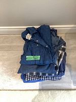 Stack of 9 folded men's shirts, including denim and plaid patterns in blue and neutral tones.