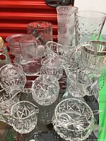 Display of assorted vintage crystal and textured glass items including covered dishes, tumblers, and vases on mirrored surface.
