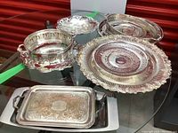 Various vintage silver-plated serving trays and bowls arranged on a glass surface showing different shapes and decorative details.