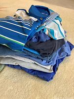 Stack of 15 men's short sleeve golf and activewear shirts in blues, striped and solid patterns, folded on carpeted floor.
