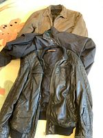 Three men's jackets in a pile on a surface: black leather jacket, brown jacket, dark synthetic jacket