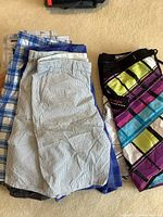Folded men's shorts laid side by side on carpet including golf and beach styles with various colors and patterns.