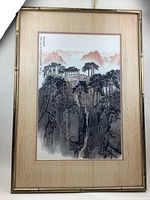Full front view of the framed Oriental landscape painting showing the mountain, trees, and building on the cliff with bamboo-style frame and beige mat with pink edging.