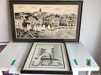 Wide shot showing both framed sketches stacked on a table, the larger coastal town sketch behind the smaller architectural sketch.