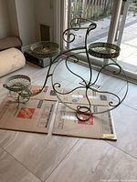 Metal garden plant stand shaped like a bicycle with three round pot holders, placed indoors on cardboard.