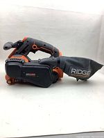 Side profile of RIDGID cordless belt sander showing handle, sanding belt housing, and dust collection bag attached with RIDGID branding.