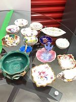 Top view of assortment of small porcelain dishes and trinket holders on glass table including oval and leaf shaped pieces with floral patterns and gold trim.