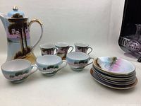 Porcelain coffee pot, cups, and saucers arranged together showing hand-painted nature scenes and gold accents.