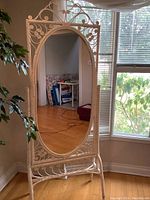 Full view of the vintage white painted wicker standing mirror situated near a window showing the oval mirror and ornate frame with stand.