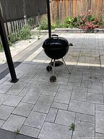 Full view of the Weber charcoal BBQ grill standing on a tiled patio. Shows the overall kettle shape, tripod stand with two wheels, and handle on lid.