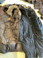 Photo showing two winter coats: a brown puffer coat with fur-lined hood labeled Parajumpers, and a black long coat with buttons and broken zipper.