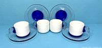 Front view of four white Guzzini espresso cups each paired with a clear glass saucer with blue circular accent underneath, arranged side by side.