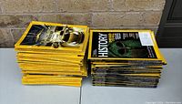Two stacks of National Geographic magazines on a table against a brick wall, approximately 64 total.