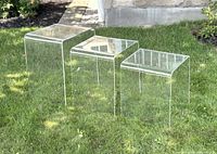 Three clear Lucite waterfall nesting tables arranged in a row on grass, visible curves and transparency showing outdoor background.