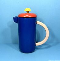 Front angle of the blue ceramic coffee maker with white handle, orange lid, and yellow knob