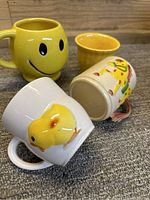 Four vintage cups highlighting the white duck Goebel cup, yellow smiley mug, horse illustrated cup, and small yellow cup on carpet