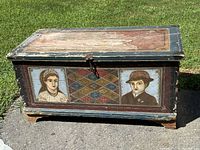 Front view of the wooden hope chest showing two painted portraits and geometric motif in between. Paint is worn and chipped revealing age.