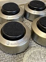 Set of four brushed metal Audio Technica acoustic feedback insulator feet with felt pads on top arranged on carpet.