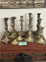 Front view showing thirteen brass candlesticks of varying heights and bases, placed on a wooden surface with a framed art piece above.