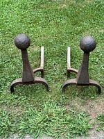 Front view of a pair of cast iron andirons with ball tops, showing rust and patina on grass background.