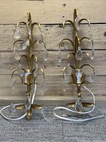 Front view of two French gilt wall sconces with hanging crystal prisms, gold-tone metal and cut electrical cords lying on the floor against wood paneling.