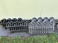 Multiple stacks of black and gray hard plastic garden fencing panels leaning against a wall outdoors on pavement with grass nearby.