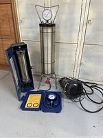 Black light leak scanner, pressure test cylinder with gauge and tripod stand, blue case with flow meter or manometer, and black cylindrical device with power cord.