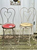 Two vintage metal ice cream parlor chairs with heart-shaped backrests, one white with a beige seat and one dark brown with a red seat. Both have chipped paint and visible wear.