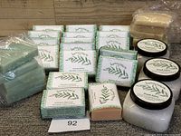 Overview of 15 wrapped soap bars with paper labels and two bags of loose soap bars, plus three jars of exfoliate scrub.