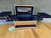 Open oak jewelry box showing black velvet interior compartments including tiered trays with multiple square sections for small jewelry items and built-in mirror.
