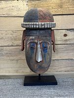 Front view of wooden Melanesian helmet mask mounted on black metal stand against wooden backdrop.
